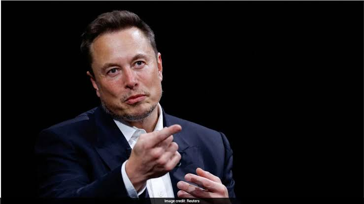 Elon Musk speaking at a press event, with SpaceX rockets in the background